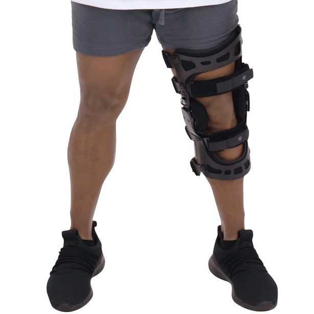 A person wearing a Vive Health 845 Dual OA Knee Brace Coretech on their left leg, standing in black sneakers and gray shorts. This black brace has adjustable straps for support, enhancing mobility and offering pain relief.