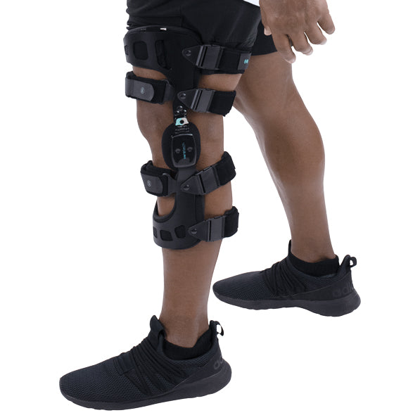 Dressed in black shorts and slip-on shoes, a person confidently stands against a white background, wearing the 845 Dual OA Knee Brace Coretech by Vive Health on the right leg. This black brace is designed for pain relief and improved mobility with multiple straps and hinges for optimal support.