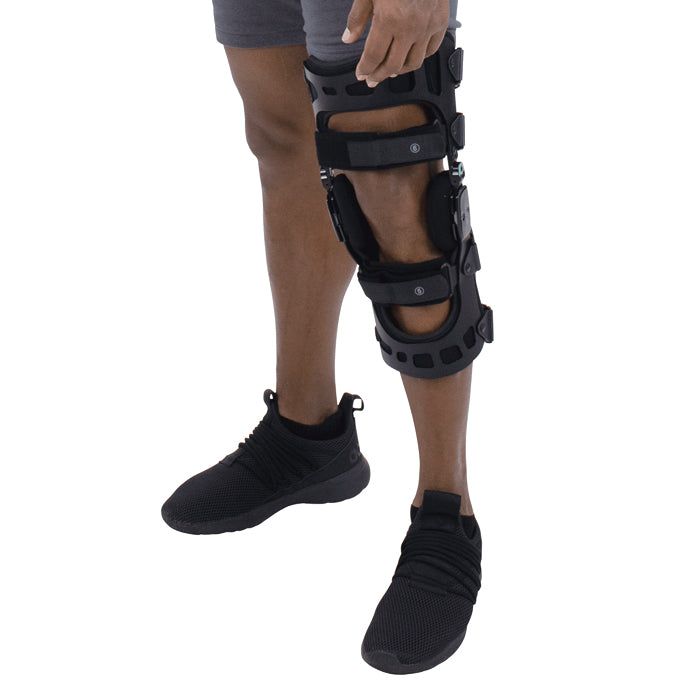 A person is wearing a Vive Health 845 Dual OA Knee Brace Coretech on their right leg for pain relief and mobility. They are dressed in dark shorts and black sneakers, while the black brace with adjustable straps provides essential knee support.