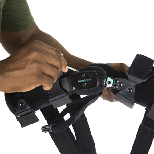 A person adjusts a Vive Health 845 Dual OA Knee Brace Coretech, a black supportive device for mobility improvement. It has mechanical components, a label, and is secured with straps. The individual skillfully operates it, possibly using a small tool.