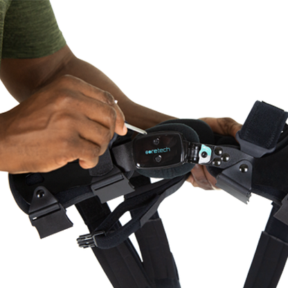 A person adjusts a Vive Health 845 Dual OA Knee Brace Coretech, a black supportive device for mobility improvement. It has mechanical components, a label, and is secured with straps. The individual skillfully operates it, possibly using a small tool.