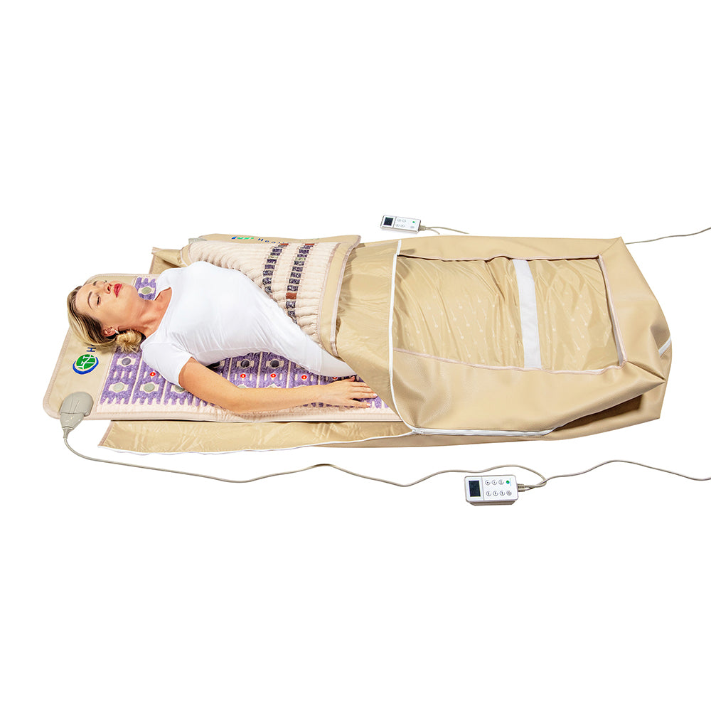 A person is enjoying a full-body experience while lying on the 360 Wrap Set™ TAJ & SOFT Full 7224 - Photon PEMF InfraMat Pro® from HealthyLine, which comes with a tan cover. The setup includes a pillow, remote control, and a transparent section over the legs. Dressed in white top and pants, they appear relaxed on the mat that features a patterned design incorporating gemstone technology for added comfort.