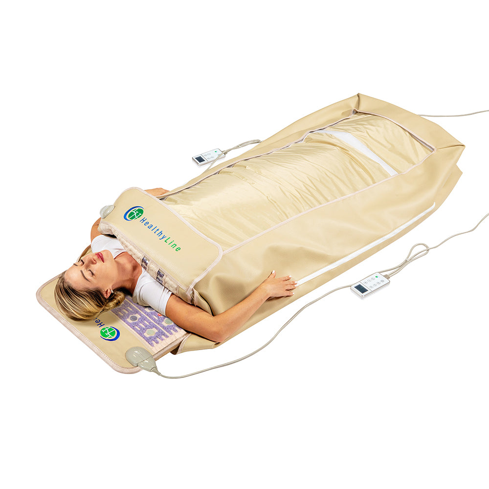 A person is lying in a beige 360 Wrap Set™ TAJ & SOFT Full 7224 by HealthyLine on a mat, enjoying soothing infrared therapy. They hold remote controls connected to the Photon PEMF InfraMat Pro®, providing a comprehensive full-body experience designed for wellness therapy.