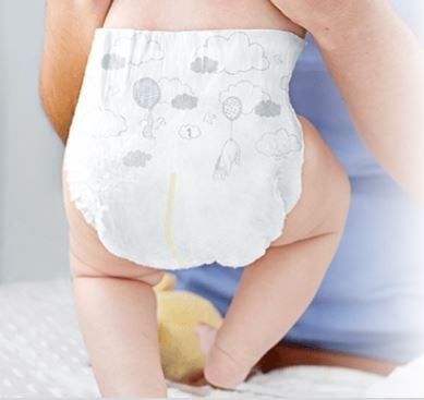 A baby in hypoallergenic McKesson Huggies Little Snugglers Baby Diapers with balloon and cloud designs sits on a lap, facing away and highlighting the gentle comfort amidst the backdrop of a blue shirt and soft textures, ensuring protection for sensitive skin.