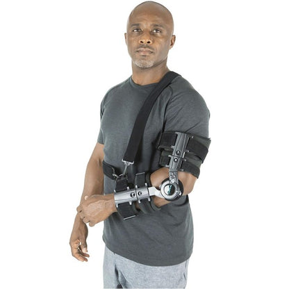 A man with a serious expression is wearing a black t-shirt and gray pants. He is equipped with the ROM Elbow Brace Coretech by Vive Health, an adjustable brace that extends from his bicep to his wrist, supported by shoulder straps for controlled range of motion. The background is a plain white studio setting.