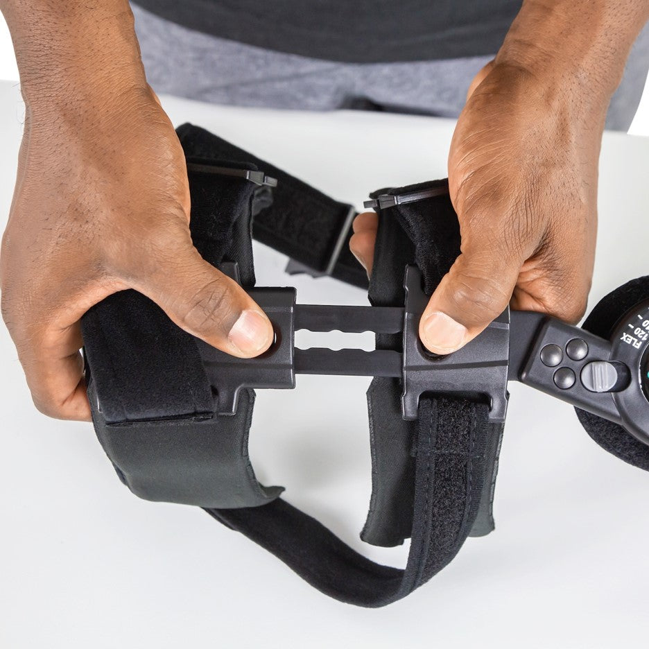 A person adjusts a ROM Elbow Brace Coretech from Vive Health, a black wearable device with adjustable straps and a central connecting part. The person's hands are demonstrating the adjustment mechanism of this supportive tool, which is designed as a rehabilitative support accessory.