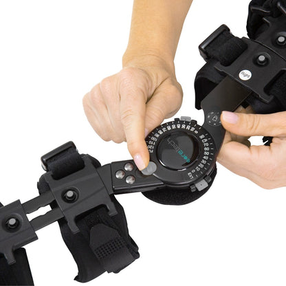 A person's hands adjust the dial on a black ROM Elbow Brace from Vive Health, featuring straps for rehabilitative support. The dial, connected to a metal hinge, precisely controls the range of motion.