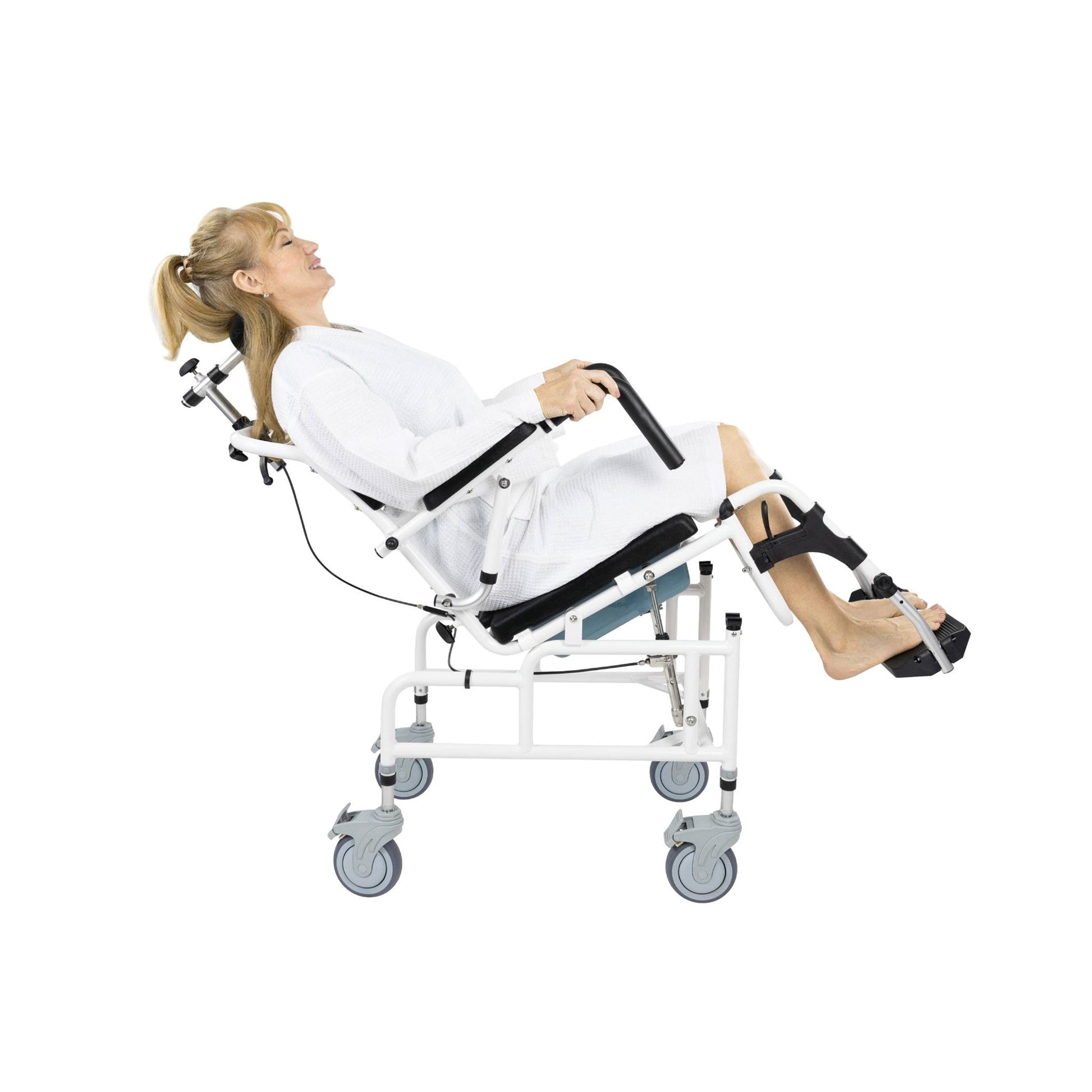 A woman in a white outfit is seated in a Vive Health Reclining Shower Commode Transport Chair, her legs elevated and supported. The sophisticated commode chair features large caster wheels and multiple adjustment mechanisms, enhancing her comfort as she relaxes and holds onto the armrests.