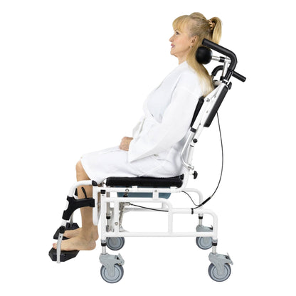 A person with long blonde hair, dressed in a white robe, sits in the Vive Health Reclining Shower Commode Transport Chair, which features leg support, armrests, and a head cushion. The person's right foot is in a cast with attached metal braces. The chair has four wheels for mobility.