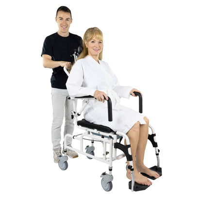 A woman sitting in a Reclining Shower Commode Transport Chair by Vive Health, smiling, and wearing a white robe. Behind her is a man standing, also smiling, and pushing the chair. The chair features armrests and footrests, and the man is holding the handles at the back.