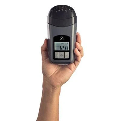 A hand holds the Breas Z2 Auto CPAP / APAP Travel Device, showcasing a reading of 12.0 on its screen. The gray device includes black and white buttons beneath the display, providing ease of use for travelers who prioritize convenience and efficiency in managing their sleep apnea while on the move.