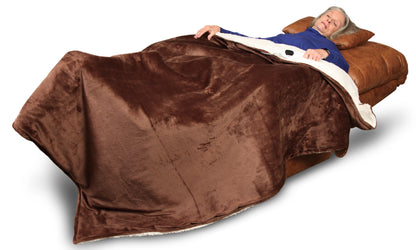 An elderly person reclines peacefully in a Perfect Sleep Chair by Journey Health & Life Style, wrapped in a large, soft brown blanket. With a relaxed expression on their face, they are dressed in a blue top and supported by a tan pillow. The dual motors provide optimal positioning beneath their head for maximum comfort.