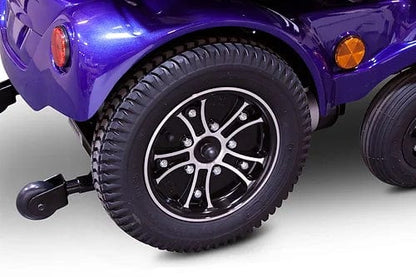 Close-up of an E-Wheels EW-M51 Power Chair's purple rear wheel shows a black tire with an intricate silver rim. Enhanced by rear-wheel drive, the smaller stabilizing wheel and orange reflectors are visible against a white background.