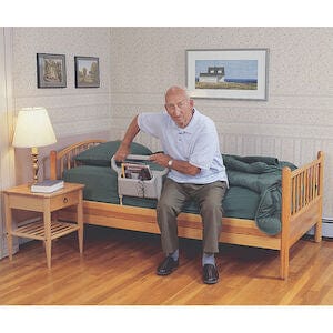 An elderly man sits on a wooden bed with a green comforter, reaching into a gray caddy filled with books. Beside him, the Bed Rail Ablerise 1EA by Maddak Inc ensures safety as he browses his collection. The cozy room features a wooden nightstand with a lamp and framed pictures adorning the walls.