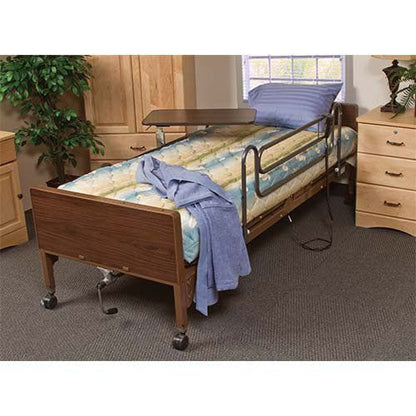 A Medline Bed Semi Electric Basic, equipped with a wooden headboard and footboard, is dressed with a blue and beige patterned blanket. A blue robe is elegantly draped over the side rail, complementing the bed's safety features that enhance comfort. Nearby, a bedside table stands against a backdrop of a window and wooden cabinets.