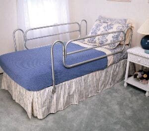 A bed adorned with blue bedding and floral pillows includes Compass Health's Bed Rails HomeStyle Carex for enhanced safety. It rests on a plush carpeted floor beside a white nightstand bearing a lamp and decorative flowers, while sheer white curtains gently drape in the background.