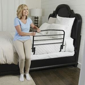 A person with shoulder-length hair and a light blue shirt is seated on a bed, holding the Stander 30" Safety Bed Rail. The room features white walls, a black headboard, and a bedside lamp.